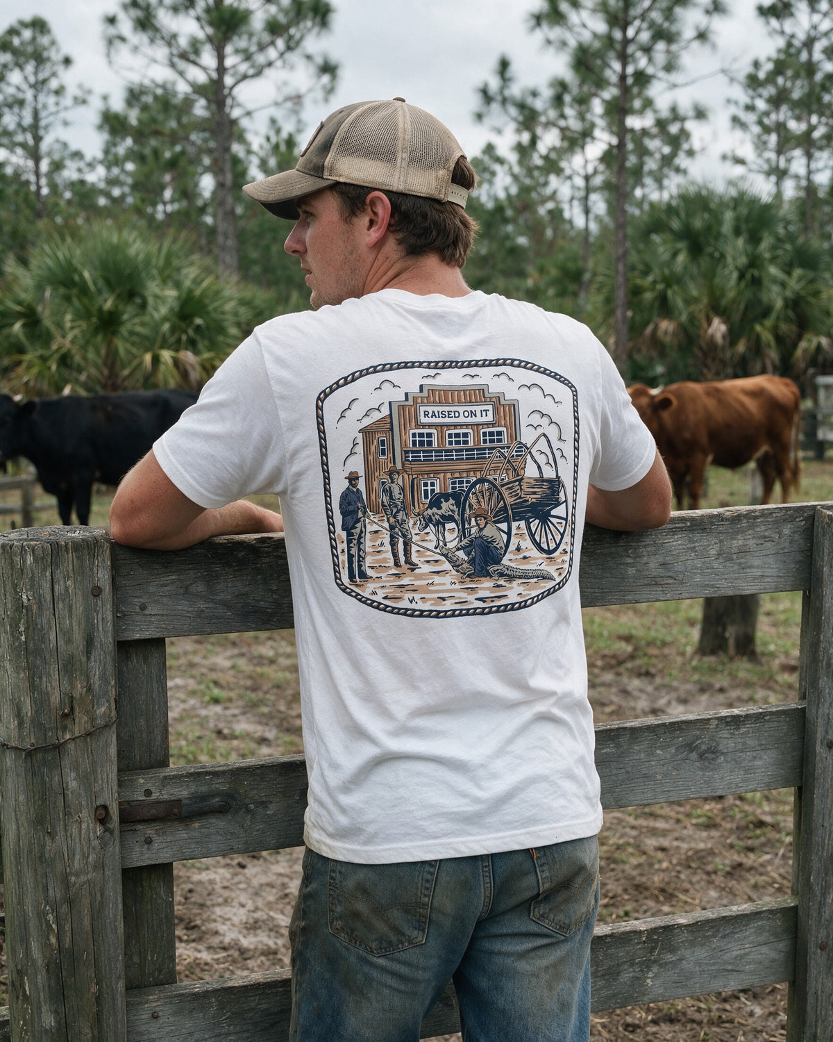 Man leaning on a cow-pen gate in the white Florida Cowboy tee, back print in view, Central Florida ranch