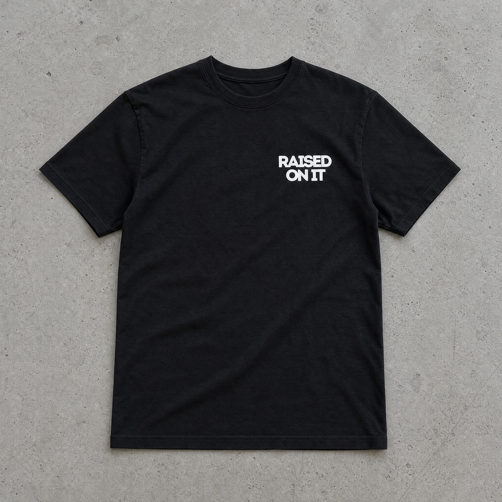 Raised On It All American Summer black tee front with chest logo, flat-lay on concrete, clean collar