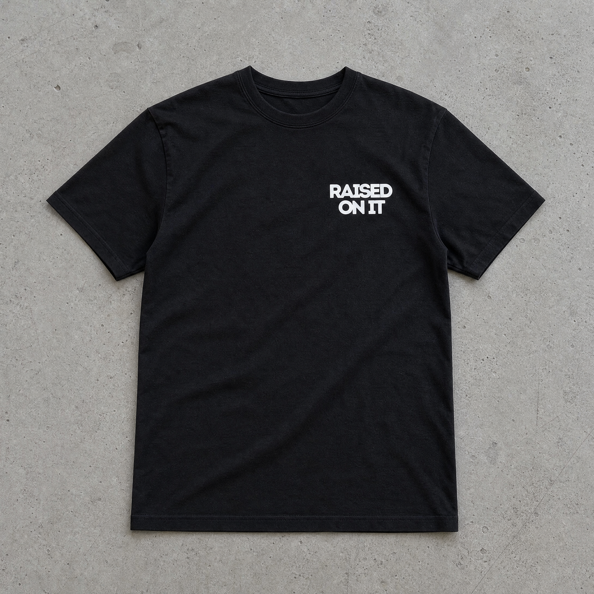 Raised On It All American Summer black tee front with chest logo, flat-lay on concrete, clean collar