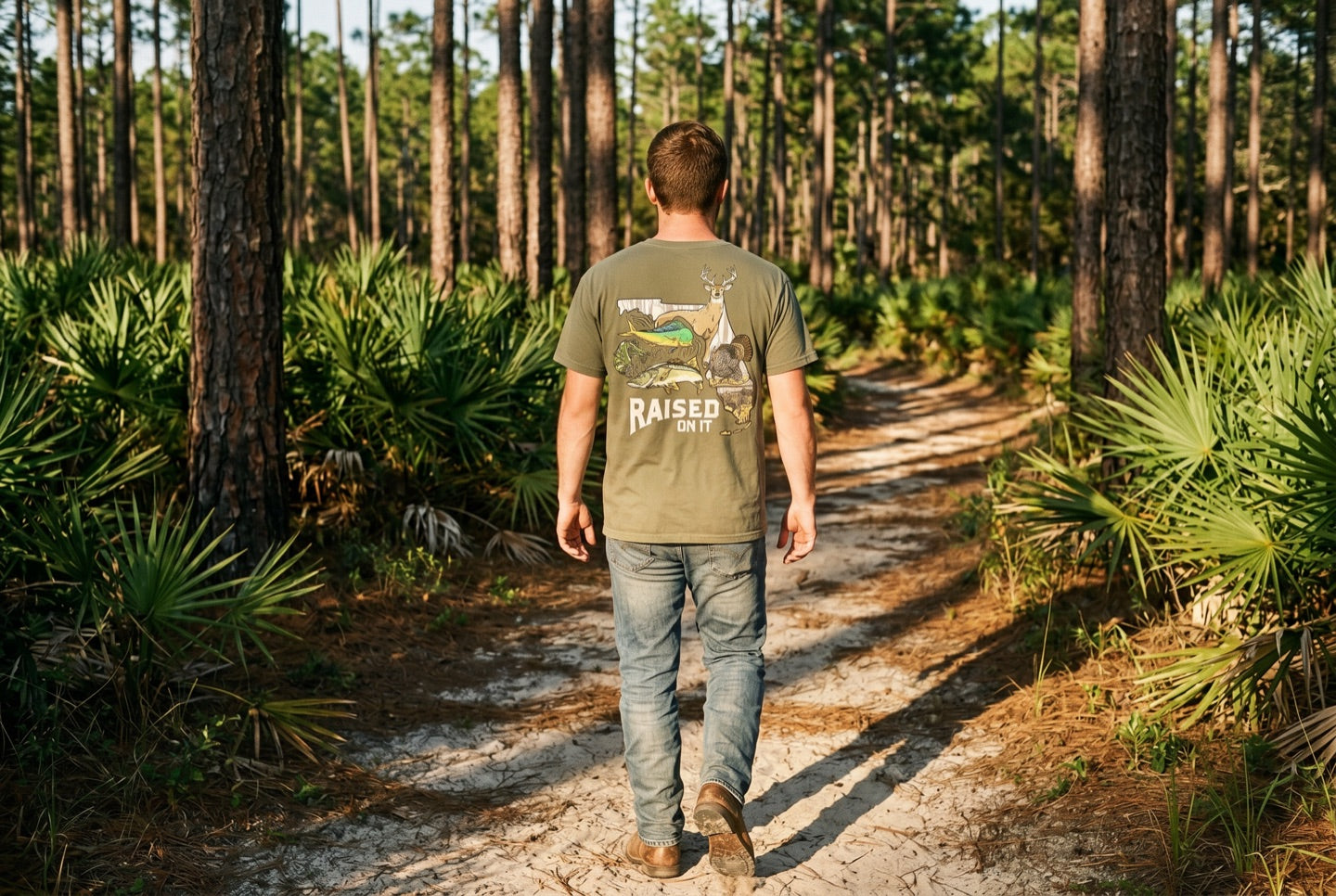 A man walking away down a Florida pine-flatwoods sand trail into golden evening light, wearing a Raised On It tee