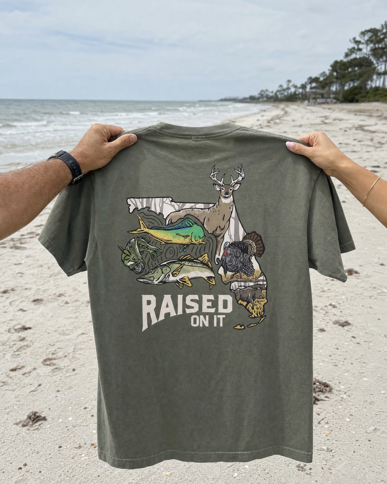 Two people holding up an olive Florida Sportsman tee on the beach